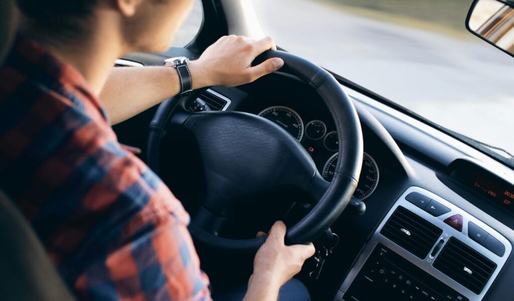 A person in a plaid shirt drives a car, both hands on the steering wheel, focused on the road ahead, with the dashboard and speedometer clearly visible.