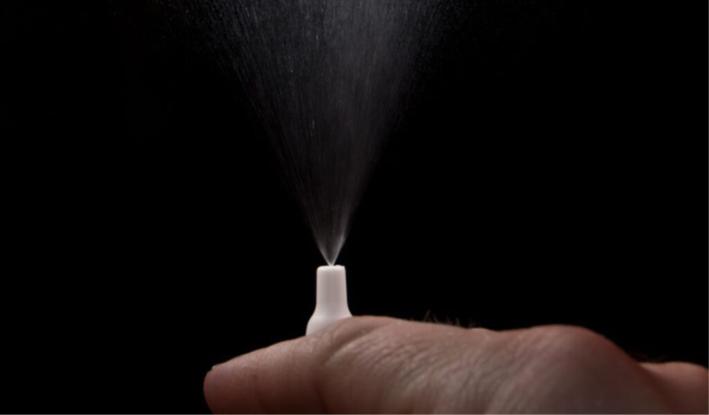 Finger pressing a small white spray nozzle, releasing a fine mist upward against a black background.
