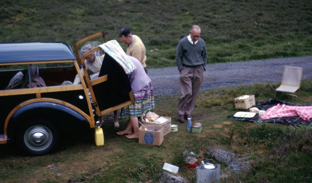 Vintage roadside camping scene with unpacked boxes, blankets, and a family beside a station wagon in a grassy area.
