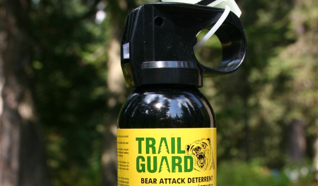 Close-up of a Trail Guard bear spray canister with safety clip intact, photographed outdoors against a blurred forest background.