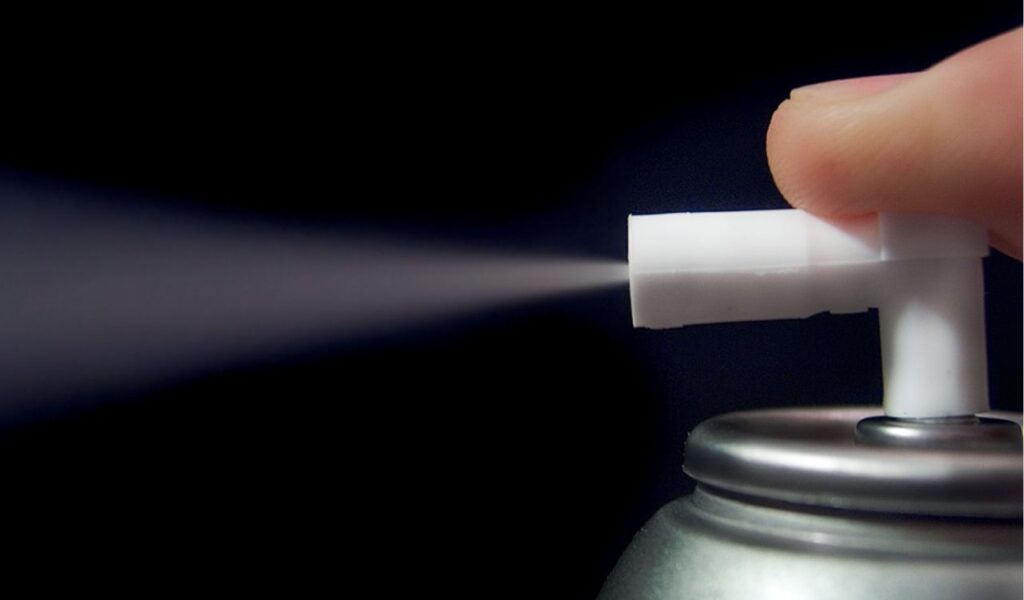 Close-up of a finger pressing a white spray nozzle, releasing mist in a dark, high-contrast background.