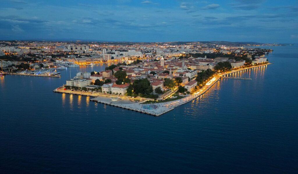 Aerial evening view of Zadar’s Old Town peninsula with glowing streetlights, historic buildings, and calm Adriatic waters reflecting the city lights.
