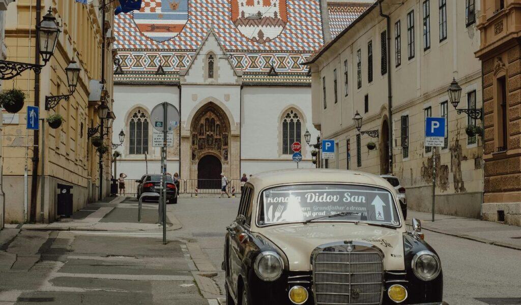 A vintage Mercedes parked on a quiet street in Zagreb's Upper Town, with the iconic tiled roof of St Mark's Church visible in the background.