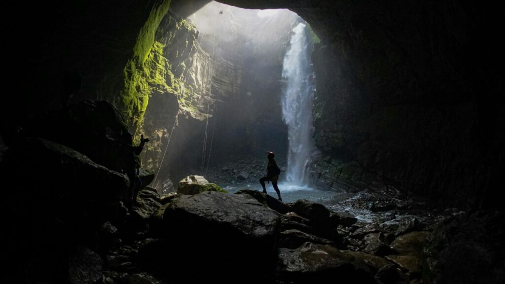 Person stands on rocks inside a large cave, looking toward a tall waterfall cascading from an opening above, with sunlight streaming in.