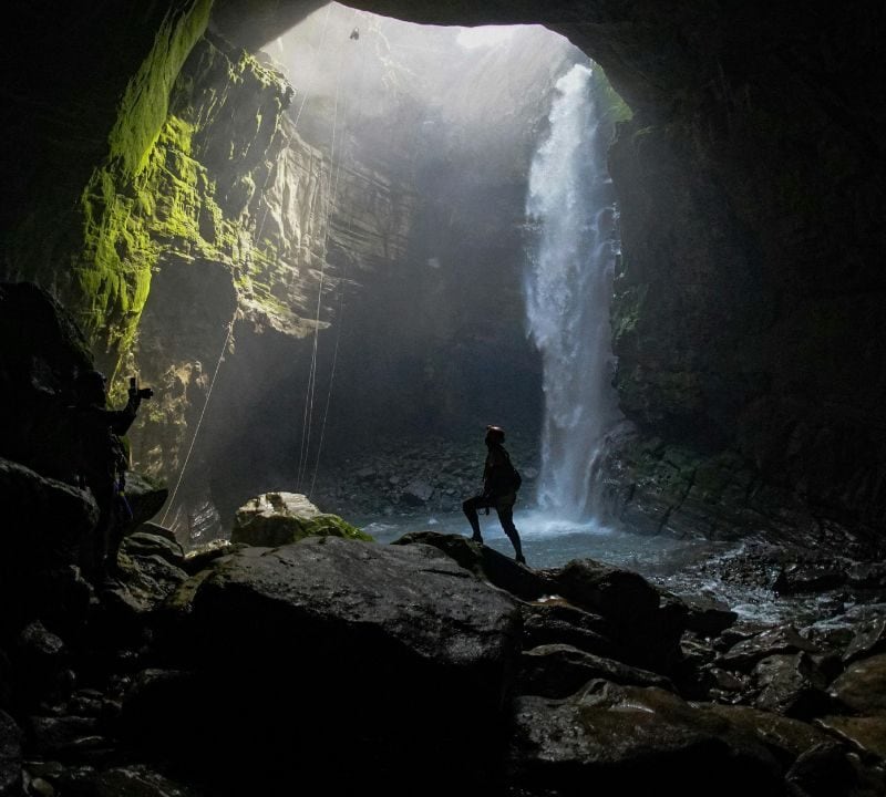 Person stands on rocks inside a large cave, looking toward a tall waterfall cascading from an opening above, with sunlight streaming in.