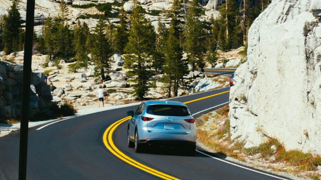 A silver car drives along a winding mountain road lined with pine trees, rocky terrain, and bright yellow road markings.