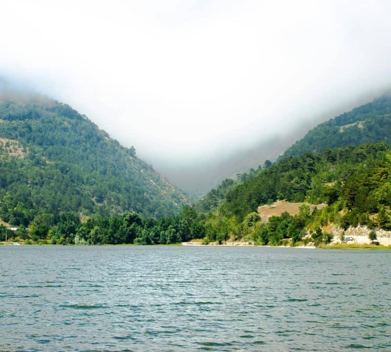 Green forested hills surrounding a calm lake, with mist and low clouds drifting through the mountain valley on a bright summer day.