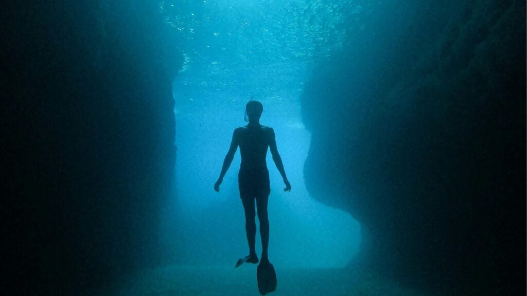 A silhouette of a diver swimming inside a deep underwater cave with rocky walls, illuminated by sunlight filtering through the water above.