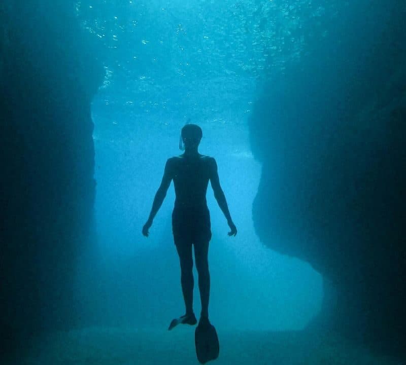 A silhouette of a diver swimming inside a deep underwater cave with rocky walls, illuminated by sunlight filtering through the water above.