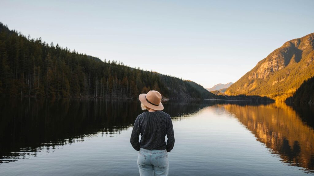 Person wearing a hat standing by a calm lake surrounded by pine forests and mountains during sunset.