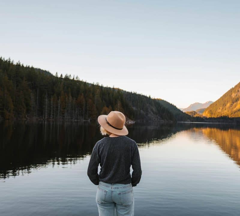 Person wearing a hat standing by a calm lake surrounded by pine forests and mountains during sunset.