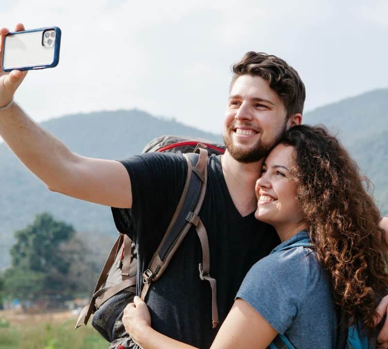 A smiling couple with backpacks takes a selfie outdoors, standing close together with mountains and greenery in the background.