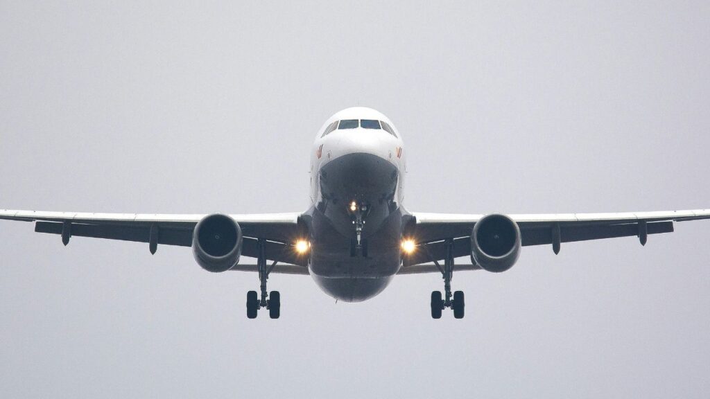A front view of a commercial airplane in flight with landing gear extended and lights on, approaching for landing against a gray sky.