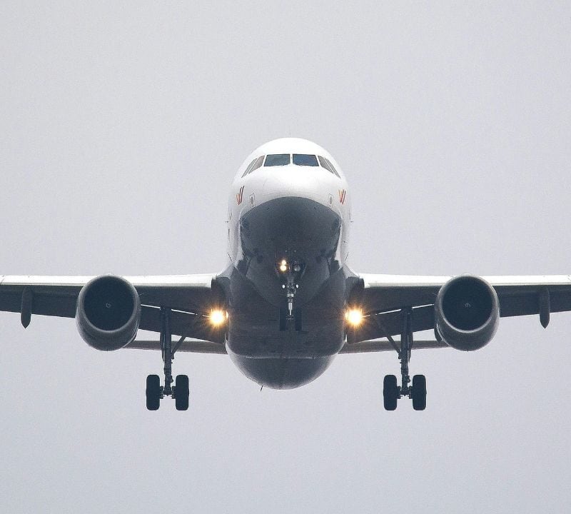 A front view of a commercial airplane in flight with landing gear extended and lights on, approaching for landing against a gray sky.
