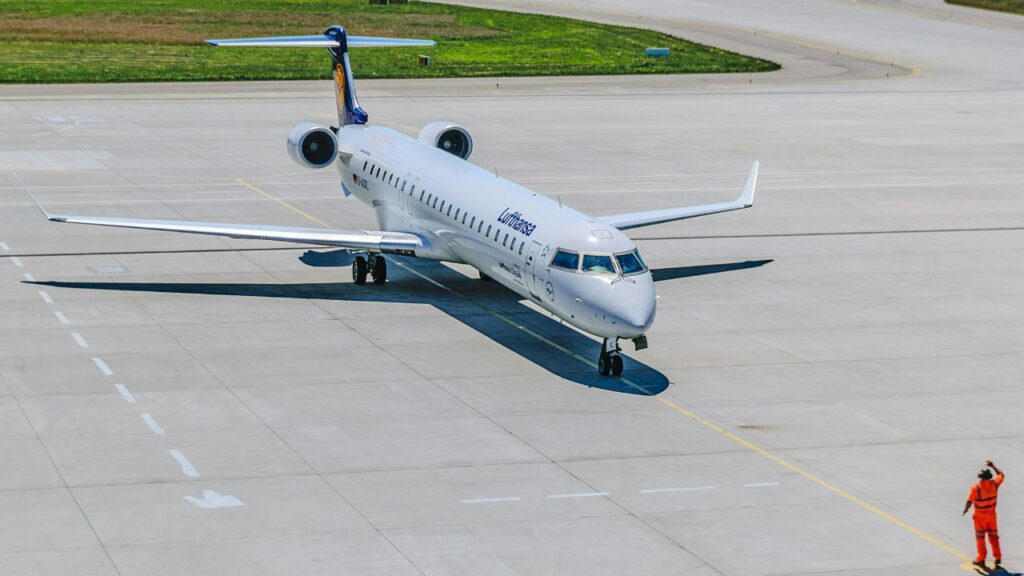 A Lufthansa airplane taxis on the runway as a ground crew member in orange directs its movement.