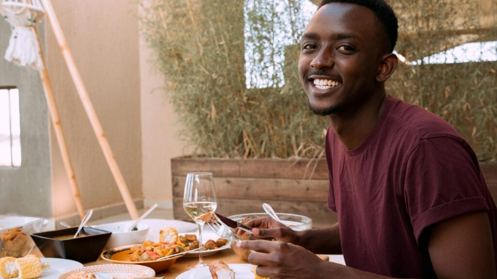 A smiling man sitting at a dining table with various dishes and a glass of wine, holding a fork and knife, ready to eat.