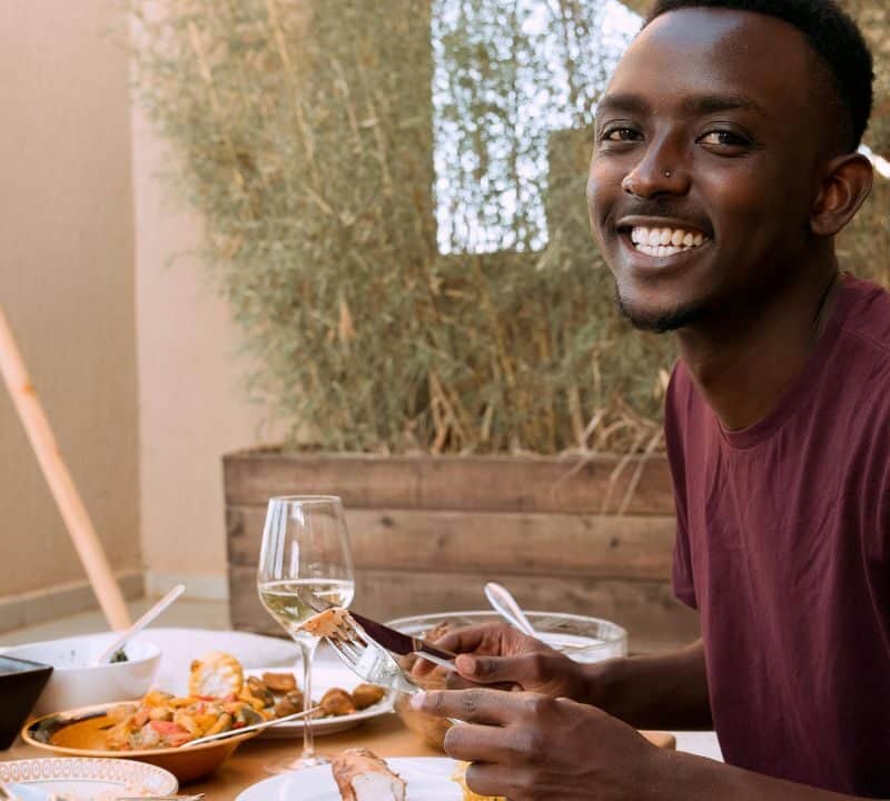 A smiling man sitting at a dining table with various dishes and a glass of wine, holding a fork and knife, ready to eat.