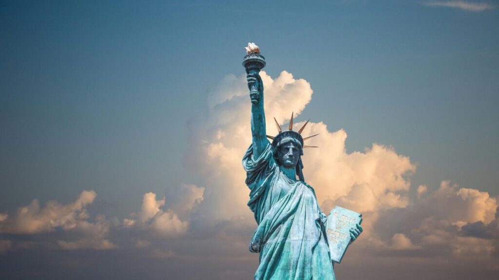 The Statue of Liberty stands tall against a backdrop of fluffy clouds and a blue sky, holding her torch high in her right hand and a tablet in her left, symbolizing freedom and democracy.