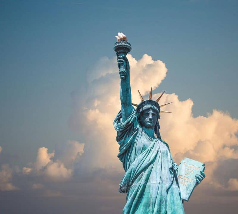The Statue of Liberty stands tall against a backdrop of fluffy clouds and a blue sky, holding her torch high in her right hand and a tablet in her left, symbolizing freedom and democracy.