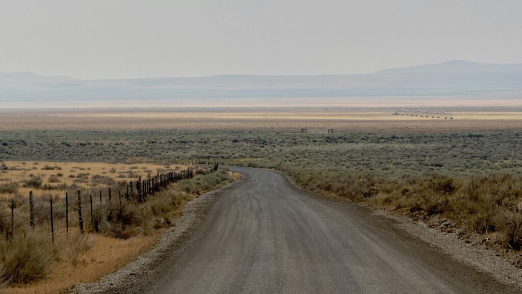 A long dirt road stretches through a dry, open landscape with sparse desert vegetation and wire fencing along one side, leading toward distant flat plains and hazy mountains under a muted sky.