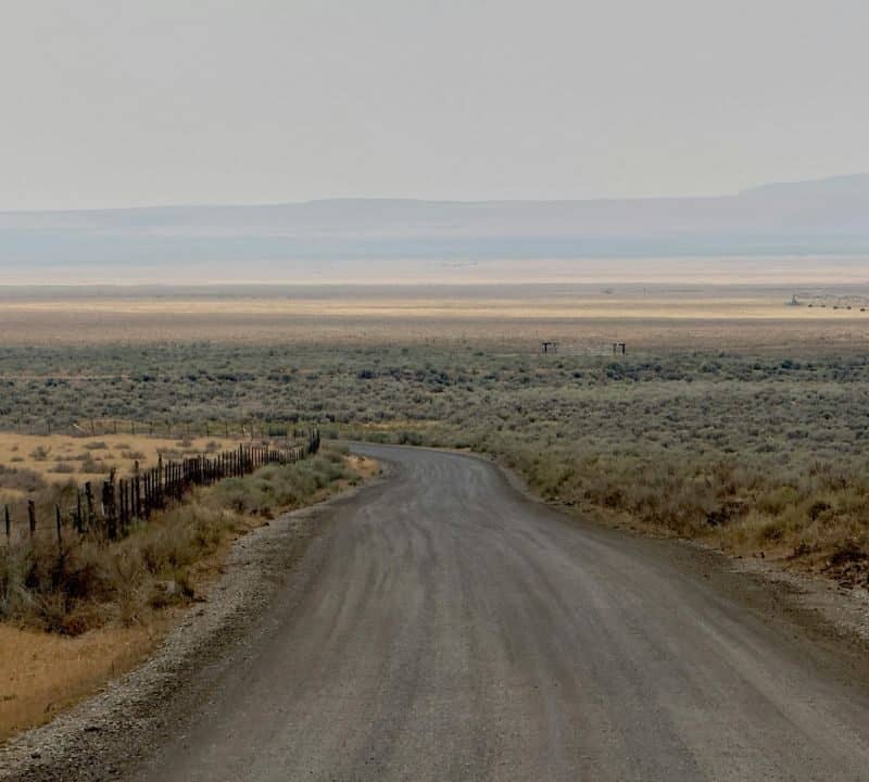 A long dirt road stretches through a dry, open landscape with sparse desert vegetation and wire fencing along one side, leading toward distant flat plains and hazy mountains under a muted sky.