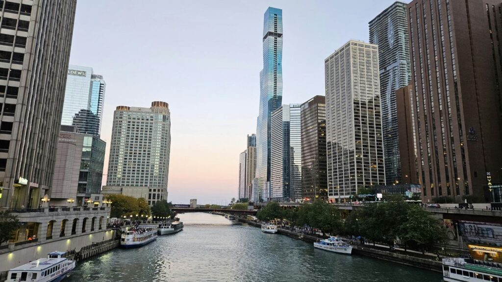 View of the Chicago Riverwalk at dusk, with tour boats along the river, modern skyscrapers on both sides, and the St. Regis Chicago tower in the background.