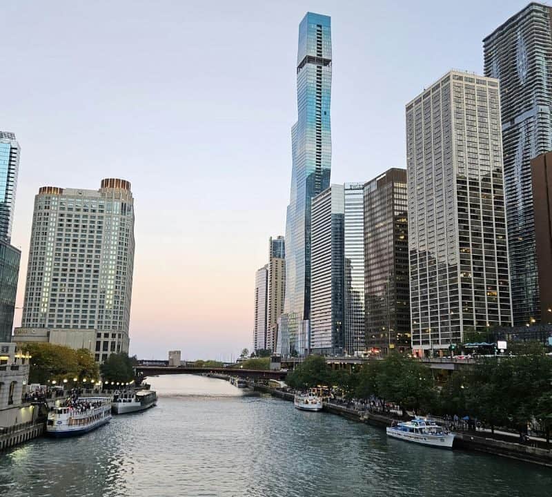 View of the Chicago Riverwalk at dusk, with tour boats along the river, modern skyscrapers on both sides, and the St. Regis Chicago tower in the background.