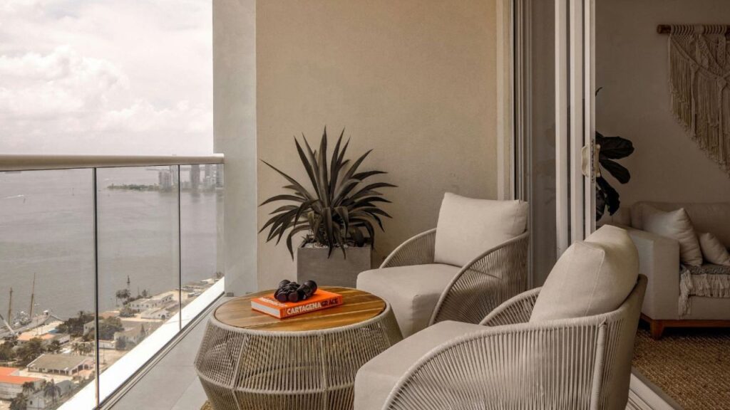 A modern Airbnb balcony setup with two cozy chairs, a small table with a book and sunglasses, a potted plant, and a view of the sea and city skyline in the distance.