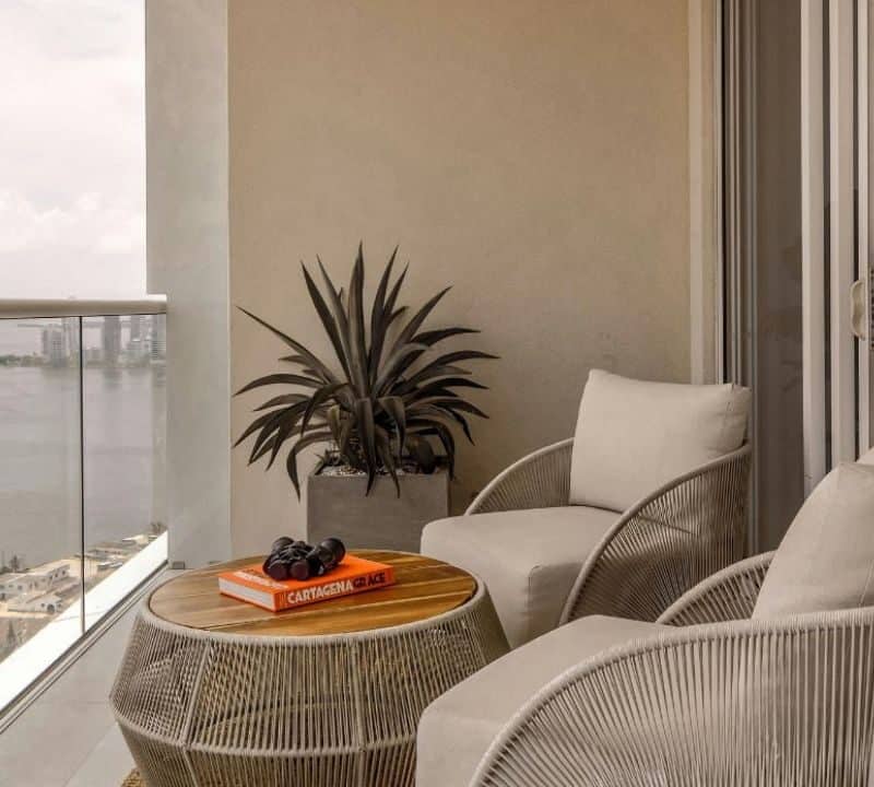A modern Airbnb balcony setup with two cozy chairs, a small table with a book and sunglasses, a potted plant, and a view of the sea and city skyline in the distance.