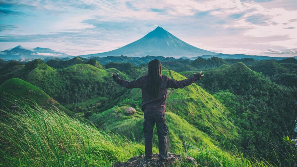 A person stands on a grassy hilltop with arms outstretched, overlooking lush green hills and a distant volcanic peak under a partly cloudy sky.