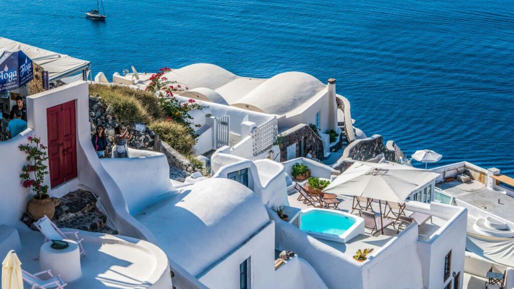 Whitewashed Cycladic buildings with curved rooftops and terraces overlook the deep blue Aegean Sea, creating a classic Greek island view.
