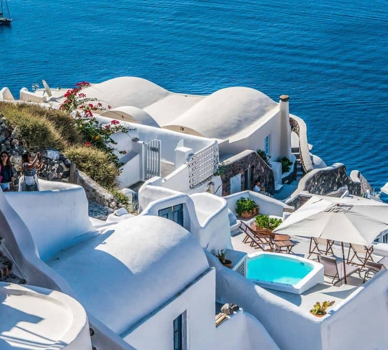 Whitewashed Cycladic buildings with curved rooftops and terraces overlook the deep blue Aegean Sea, creating a classic Greek island view.