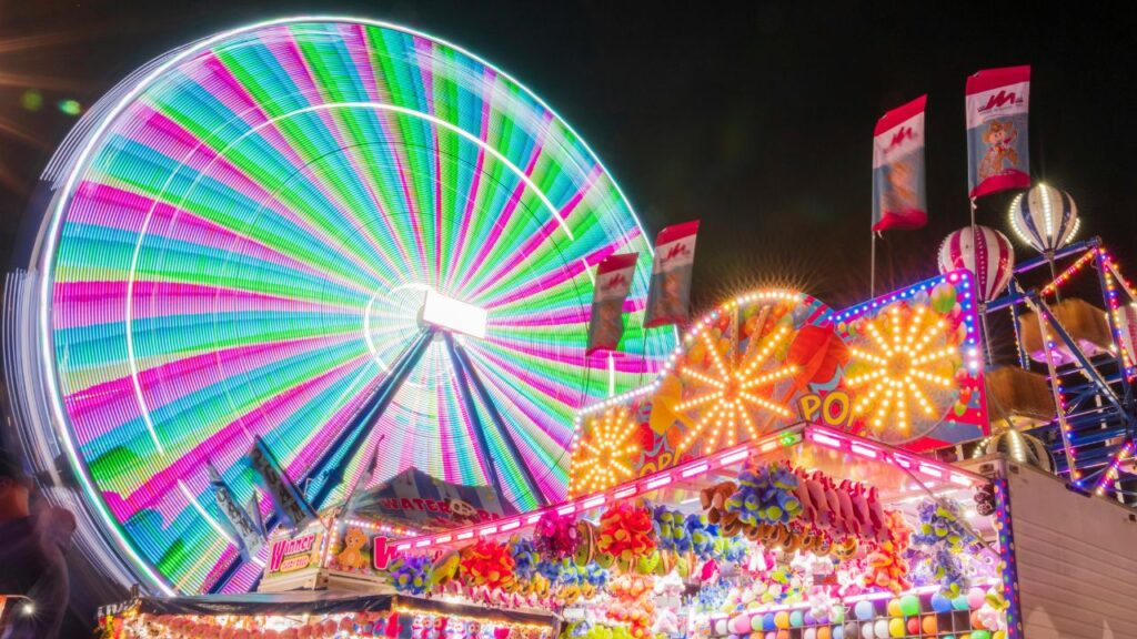 A brightly lit Ferris wheel glows in rainbow colors at night beside a carnival game booth filled with plush toys and flashing lights.