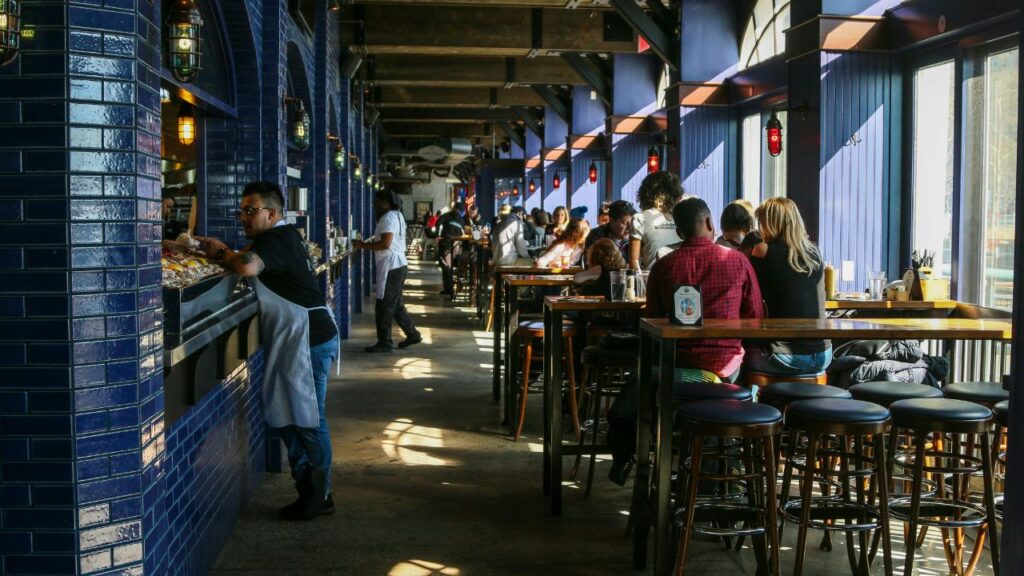 A busy indoor restaurant with deep blue tiled walls and high wooden tables, where diners enjoy meals and staff work behind a long counter.