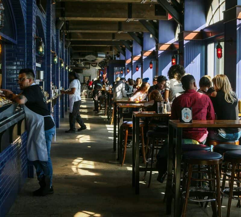 A busy indoor restaurant with deep blue tiled walls and high wooden tables, where diners enjoy meals and staff work behind a long counter.