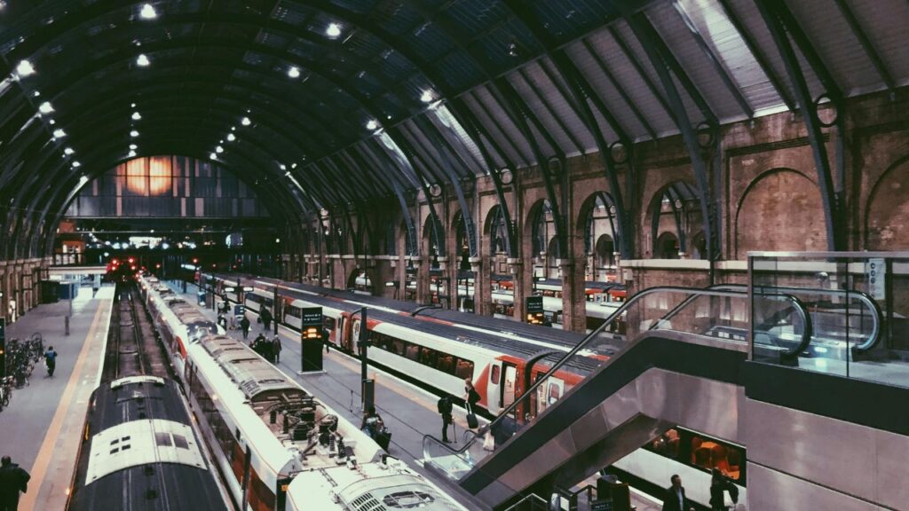 Interior view of a busy London train station with multiple platforms, modern trains, and passengers walking under a high arched roof.