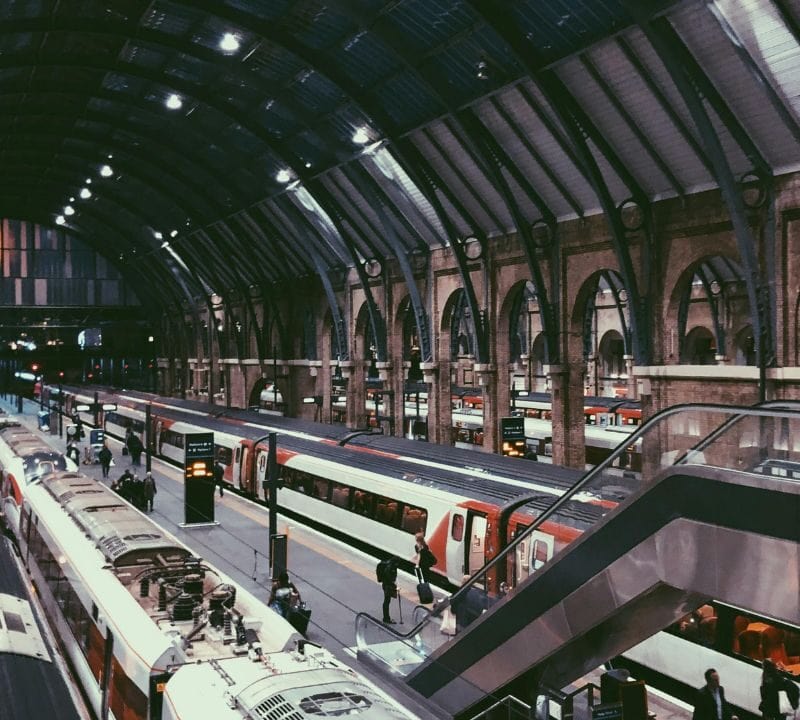 Interior view of a busy London train station with multiple platforms, modern trains, and passengers walking under a high arched roof.