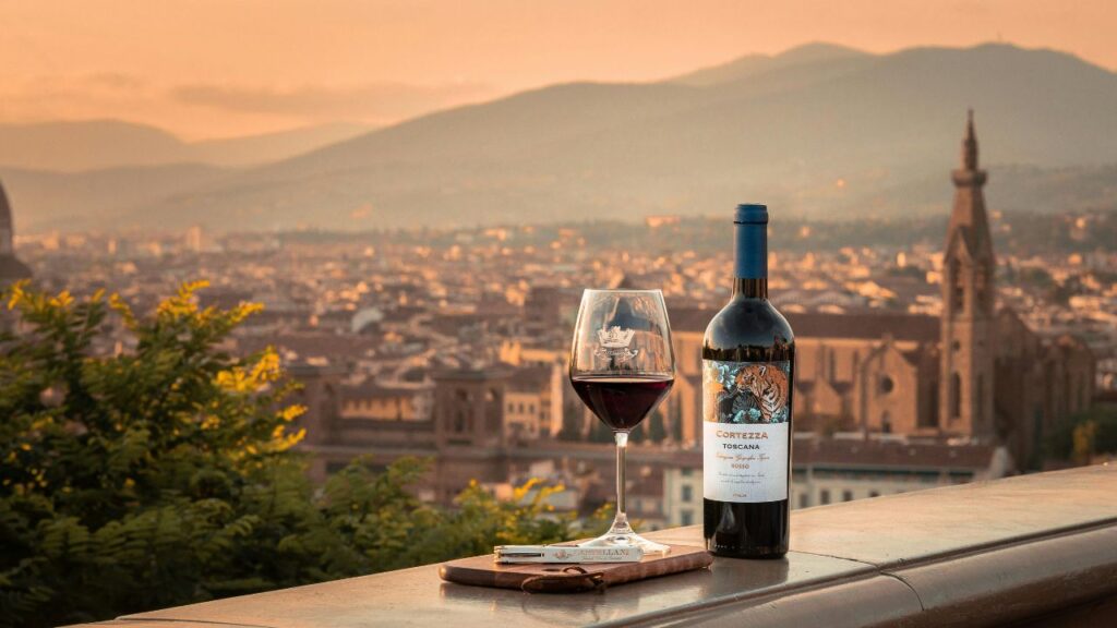 A bottle of Tuscan red wine and a filled glass sit on a ledge overlooking Florence at sunset, with the city’s rooftops and mountains in the distance.