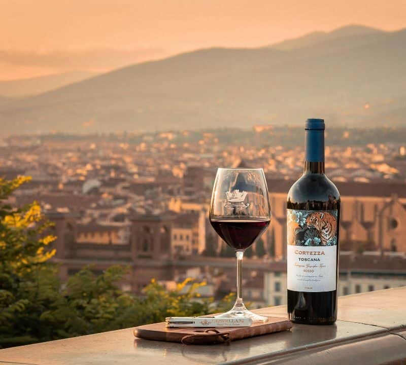 A bottle of Tuscan red wine and a filled glass sit on a ledge overlooking Florence at sunset, with the city’s rooftops and mountains in the distance.
