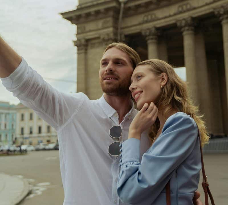 A couple taking a selfie together in front of a grand building with tall columns, smiling and enjoying their travel moment.