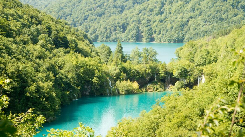 A turquoise lake surrounded by dense green forested hills, with small waterfalls cascading into the water, creating a serene and pristine natural scene.