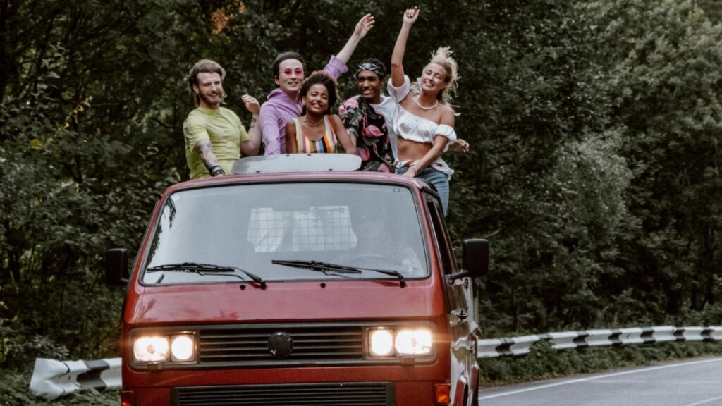 Five friends stand through a red van's sunroof, smiling and raising arms while driving on a tree-lined winding road.