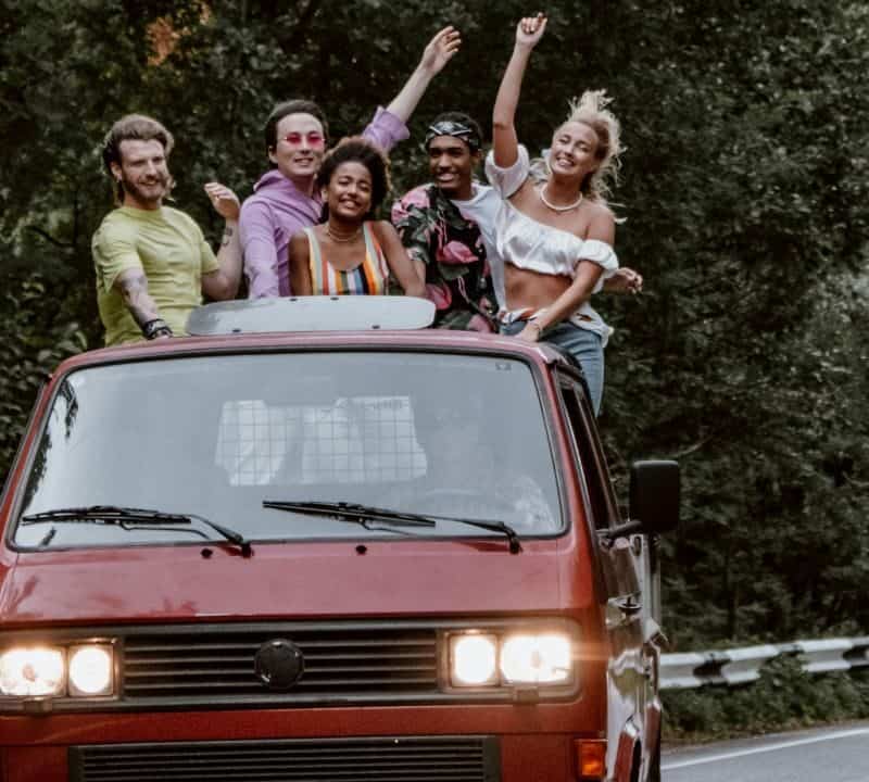 Five friends stand through a red van's sunroof, smiling and raising arms while driving on a tree-lined winding road.