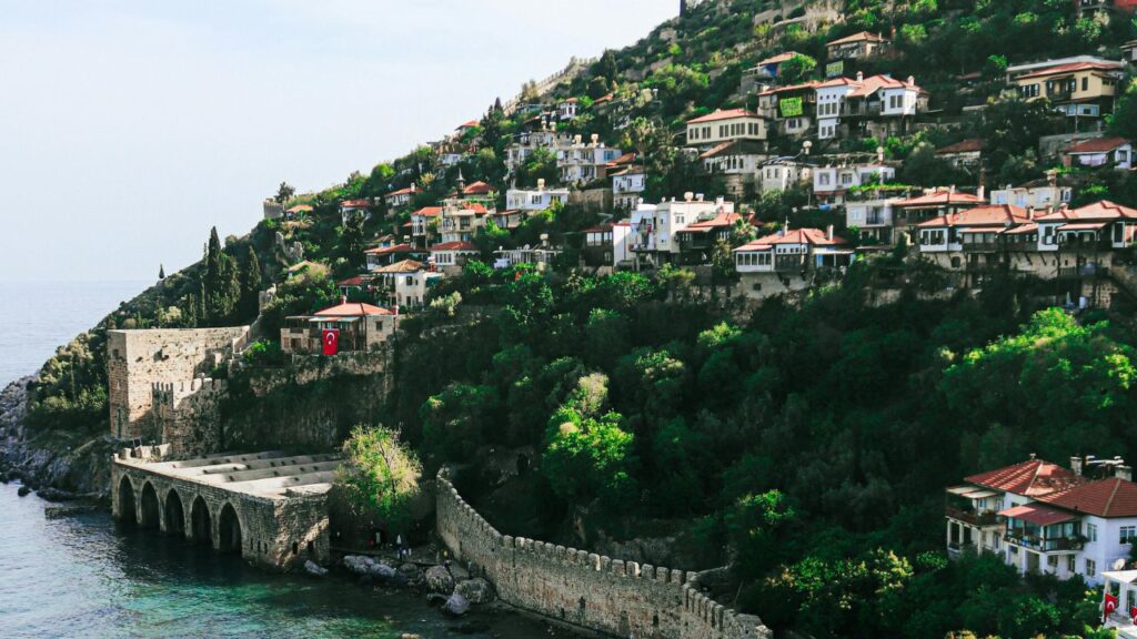 Colorful hillside homes overlook the sea in Alanya, Turkey, with a historic stone dockyard and fortress wall along the turquoise coastline.