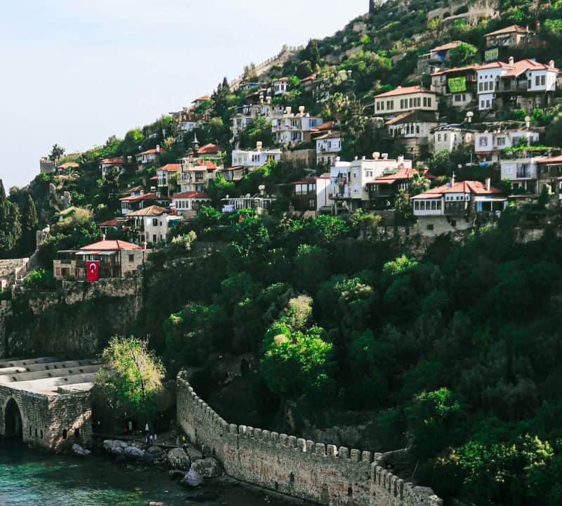Colorful hillside homes overlook the sea in Alanya, Turkey, with a historic stone dockyard and fortress wall along the turquoise coastline.