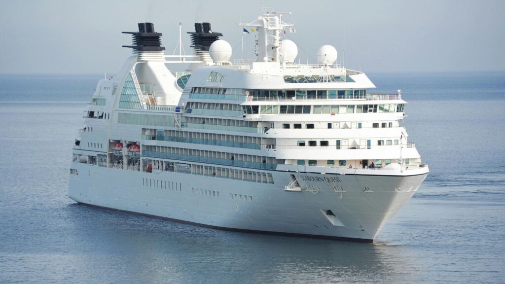 A large white luxury cruise ship, Seabourn Quest, sails on calm blue waters with multiple decks, balconies, and radar domes visible.