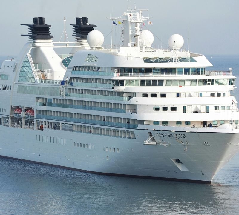 A large white luxury cruise ship, Seabourn Quest, sails on calm blue waters with multiple decks, balconies, and radar domes visible.