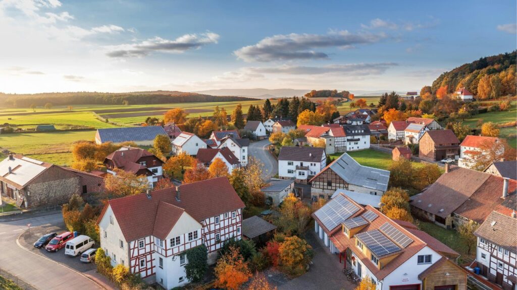 Aerial view of a small countryside village with red-roofed houses, solar panels, winding streets, and surrounding green and autumn-colored fields under a partly cloudy sky.