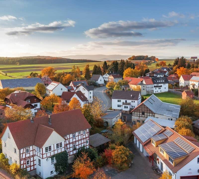 Aerial view of a small countryside village with red-roofed houses, solar panels, winding streets, and surrounding green and autumn-colored fields under a partly cloudy sky.