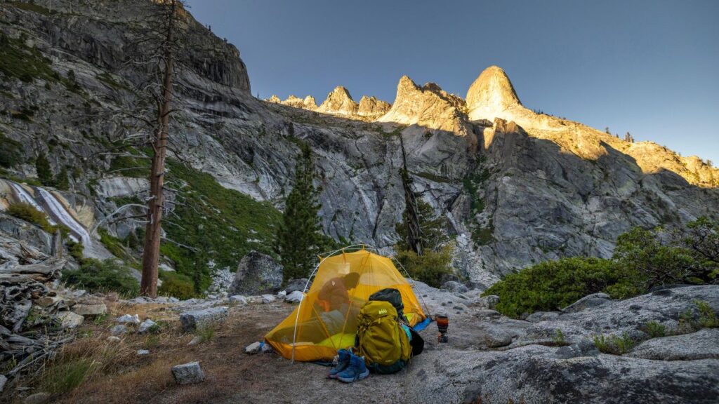  A bright yellow tent set up on rocky terrain in the mountains, with a backpack and hiking boots outside, surrounded by steep granite cliffs and sunlit peaks in the background.